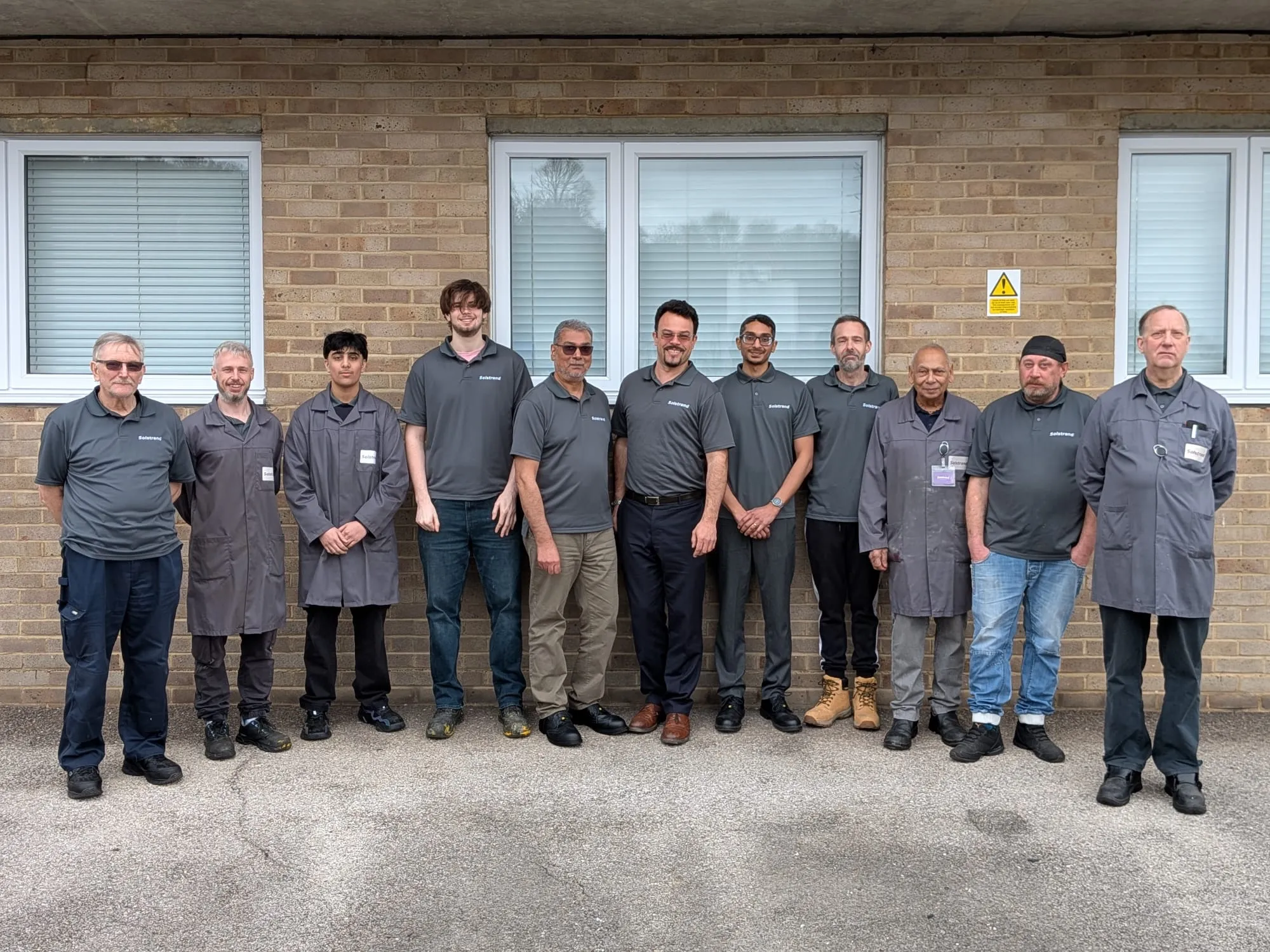 Eleven members of the Solstrand team standing in a line outside a brick building, wearing grey company polo shirts and work coats