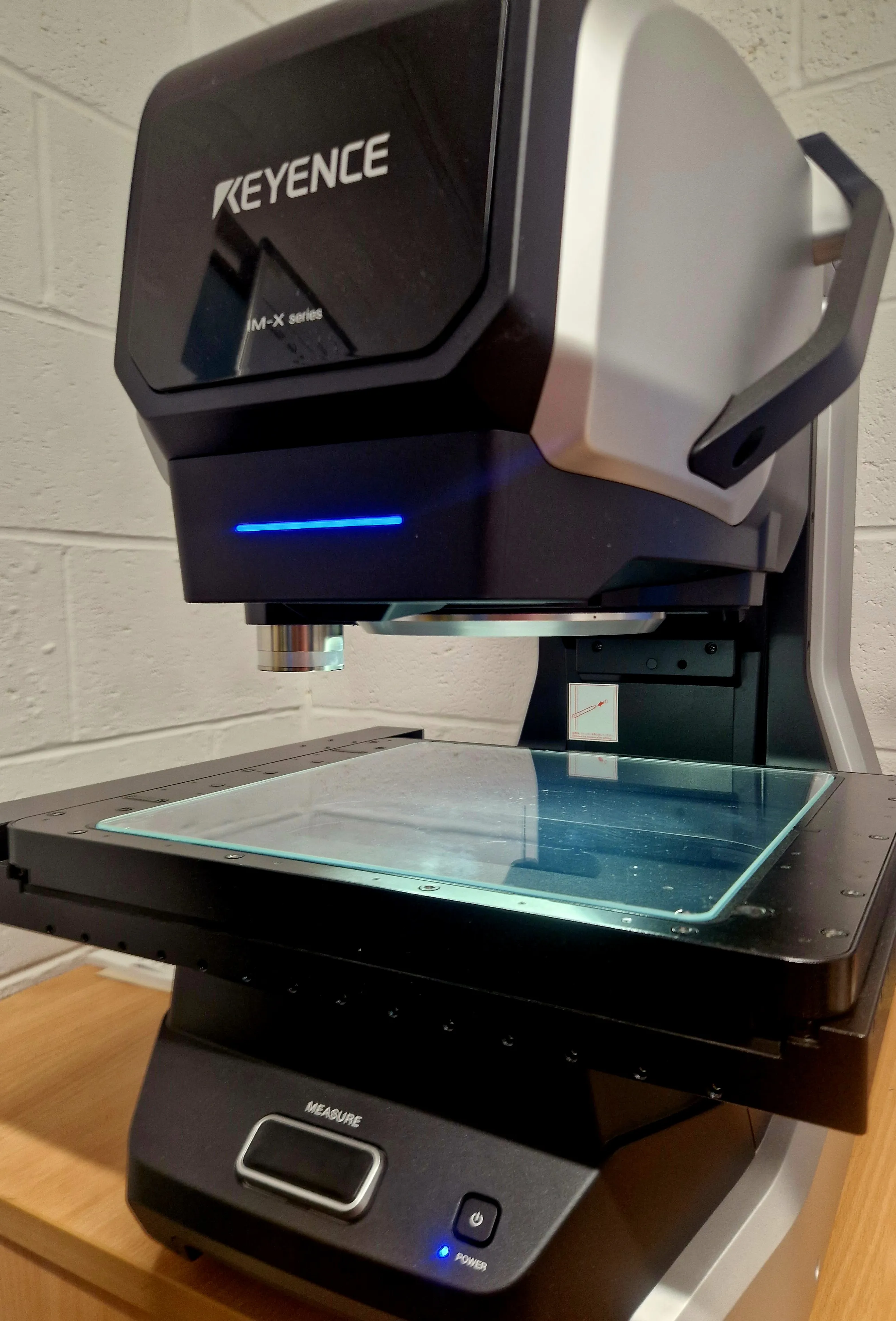 Close-up of the Keyence IM-X1000 measurement system, showing its glass measuring stage and objective lens
