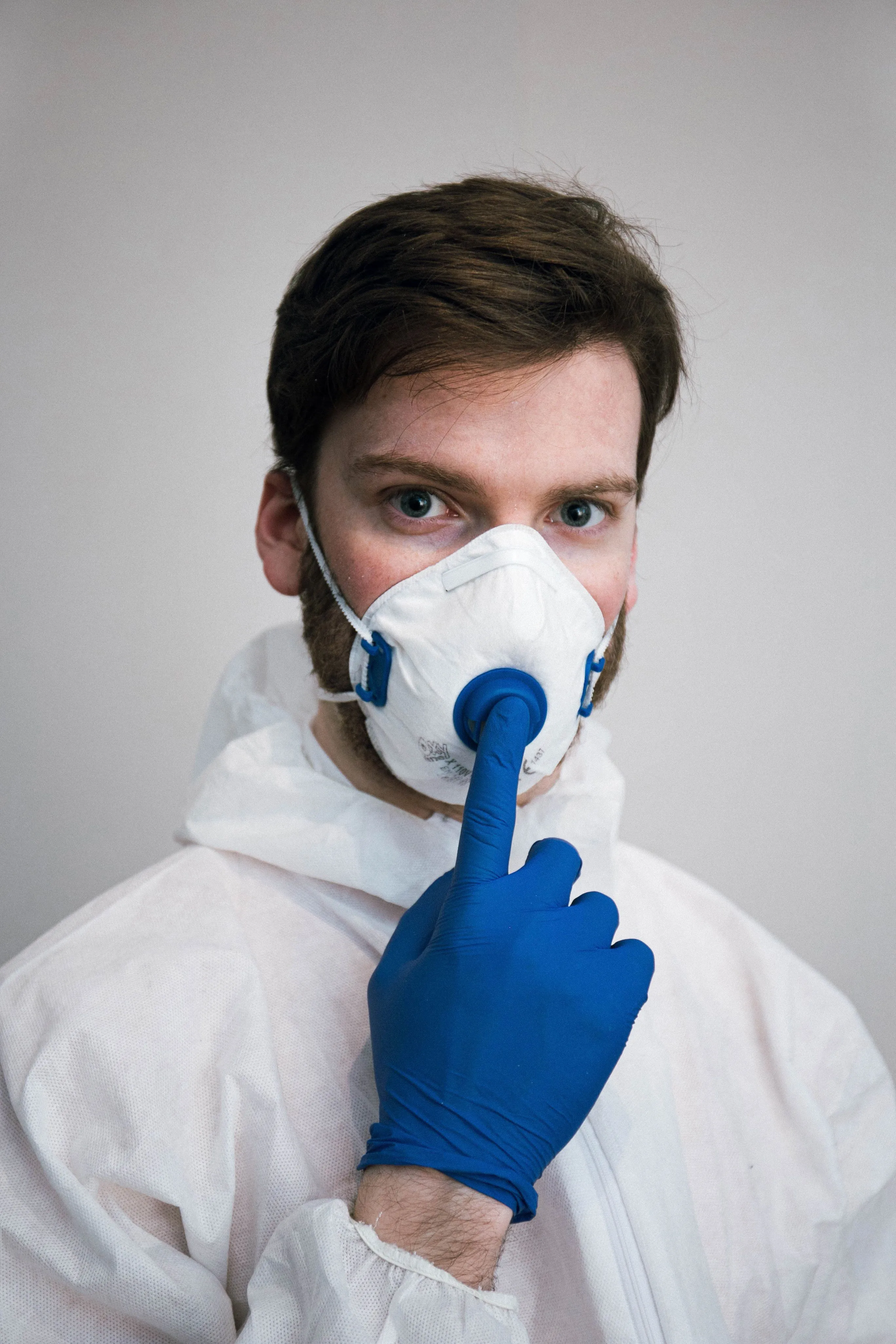 A person in a white protective suit and blue glove, testing the fit of a white respirator mask